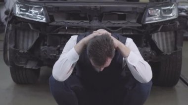 Kırık arabanın önünde oturan ve el ele tutuşan sakallı beyaz adamın portresi. Bozuk otomobilin arka planındaki umutsuz iş adamı. Araba kazası, tamirhane, garanti servisi..