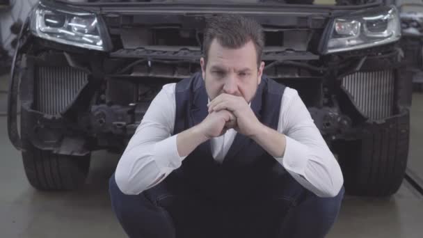 Portrait of desperate Caucasian man sitting in front of broken car and holding head with hands. Upset adult businessman at the background of broken automobile. Crash, car accident, repair shop. — 비디오