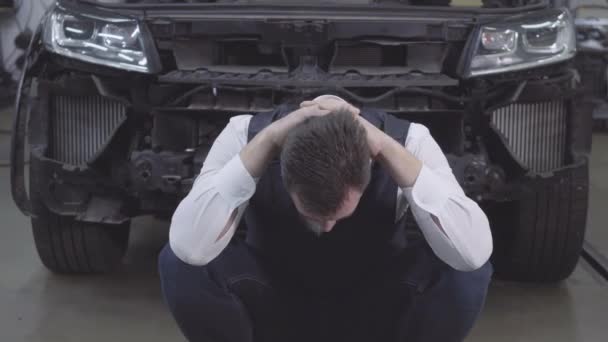 Portrét vousatého bělocha sedícího před rozbitým autem a držícího se za ruce. Zoufalý obchodník na pozadí rozbitého auta. Autonehoda, opravna, záruční servis. — Stock video