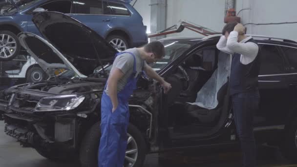 Uomo adulto caucasico in piedi accanto alla sua macchina rotta e tenendo la testa con le mani. Meccanico automatico in abito da lavoro esaminando rotto automobile con il cliente. Incidente stradale, servizio di riparazione . — Video Stock