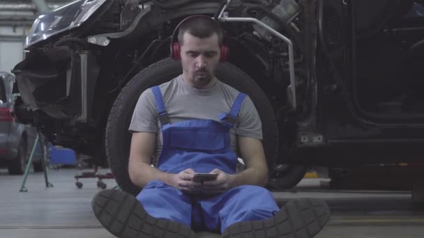 Portrait d'un mécanicien automobile caucasien fatigué s'appuyant sur la roue de la voiture et écoutant de la musique dans les écouteurs. Professionnel épuisé prenant une pause sur le lieu de travail. Mode de vie, travail, profession . — Video
