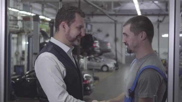 Side view close-up of two Caucasian men shaking hands in repair shop and talking. Auto mechanic and client standing at the background of cars. Workshop, warranty service. — 비디오