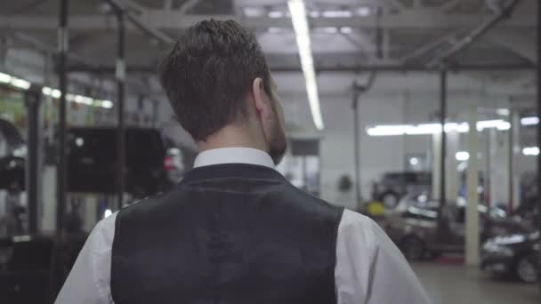 Vista posteriore dell'uomo caucasico in giubbotto e camicia in piedi in officina e guardando le auto. Giovane uomo d'affari fiducioso in attesa che la sua auto venga riparata. Garanzia, servizio, riparazione auto . — Video Stock