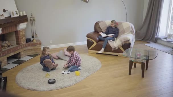 Long plan de trois enfants caucasiens d'âges différents assis à la maison devant la cheminée le week-end. Jeunes enfants jouant sur un tapis mou, sœur aînée assise sur un canapé et lisant un livre . — Video