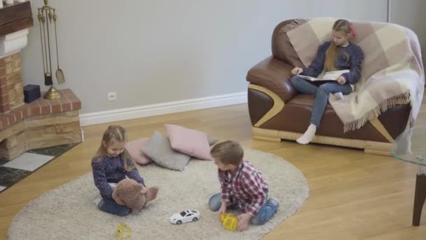 Top view of cheerful Caucasian brother and sister playing at home as their elder sister reading book. Calm children spending free time together at home. Leisure, lifestyle. — Stock Video
