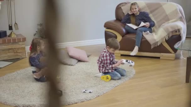 Tre vita barn sitter hemma och pratar. Liten flicka och pojke som håller leksaker som sin tonårssyster sitter på soffan med bok. Barn vilar inomhus på helgerna. — Stockvideo
