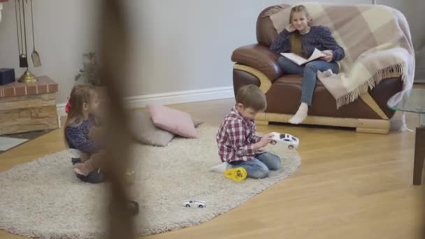 Three Caucasian children sitting at home and talking. Little girl and boy holding toys as their teenage sister siting on sofa with book. Kids resting indoors on weekends. — Stock Video