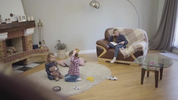 Porträt eines süßen kaukasischen Mädchens, das auf einer Couch mit Buch sitzt, während ihre jüngere Schwester und ihr Bruder auf dem Boden spielen. Ältere Geschwister kümmern sich zu Hause um kleine Kinder. Glück, Einheit, Familie. — Stockvideo
