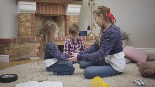 Joyful Caucasian girl teaching younger brother and sister playing a game. Happy teenager spending free time with kids at home. Unity, happiness, leisure.