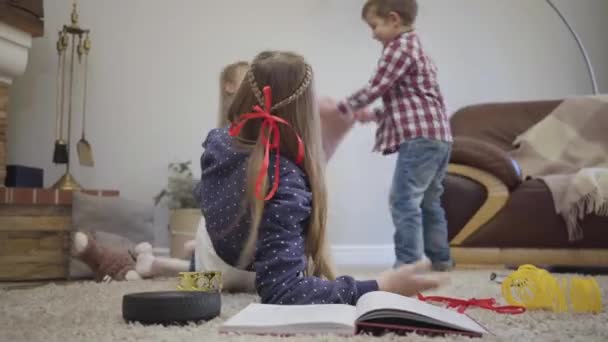 Close-up of teen Caucasian girl turning back and talking to younger brother and sister fighting with pillows. Little siblings distracting teenager from education. Leisure, unity, lifestyle. — 비디오