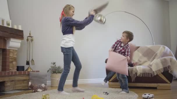 Side view of Caucasian girl in casual clothes fighting with pillows with younger siblings. Happy girls and boy playing together at home. Leisure, lifestyle, happiness. — 비디오