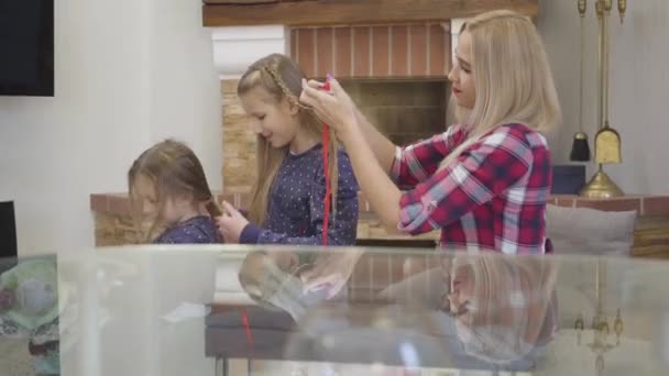 Vista lateral de la madre rubia caucásica y dos hijas trenzándose el pelo. Personas reflejándose en la mesa de vidrio de pie en primer plano. Felicidad, unidad, ocio . — Vídeos de Stock