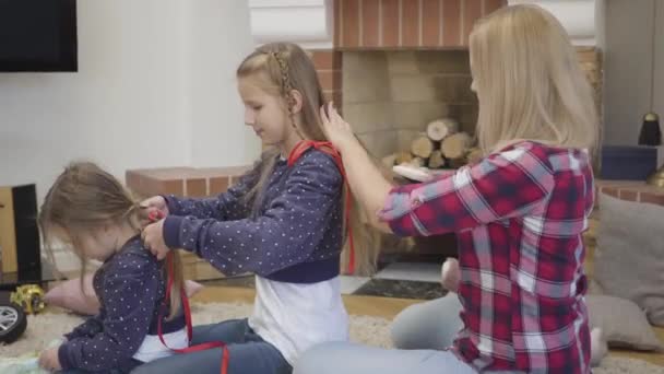 Вид сбоку на белокурую белую женщину, расчесывающую волосы старших дочерей, когда она плетёт косички для младшей сестры. Мама проводит свободное время с детьми в помещении. Счастье, единство, досуг . — стоковое видео