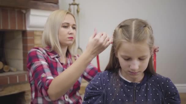 Camera uitzoomen. Blond volwassen blanke vrouw doet haarjurk voor oudere dochter, tienermeisje vlechten jongere zus, kleine schattige dame kammen pop. Zorg, levensstijl, vrouwelijkheid. — Stockvideo