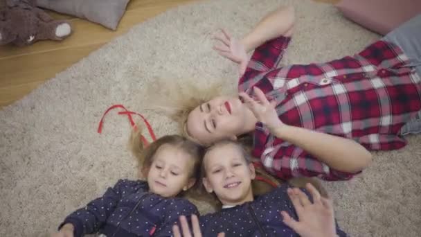 Камера наближається до молодої кавказької матері, яка лежить з дочками на м'якому килимі, тримаючись за руки. Весела сім'я відпочиває вдома на вихідних. Щастя, єдність, відпочинок. — стокове відео