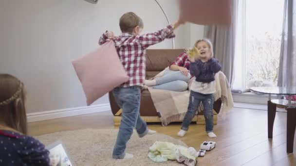 Graciosos niños caucásicos peleando con almohadas y corriendo como su madre sentada en el sofá y su hermana mayor tomando selfie. Feliz hermano y hermana divirtiéndose con la familia en casa . — Vídeos de Stock