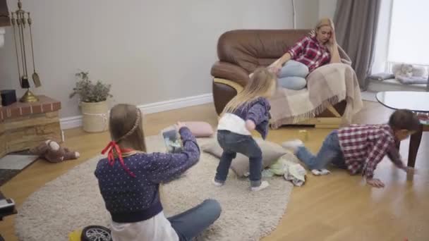 Müde Kaukasierin, die auf dem Sofa ruht, während ihre Kinder spielen. Der kleine Sohn und die kleine Tochter laufen als ältere Mädchen mit dem Tablet für ein Selfie herum. große Familie, Glück der Mutterschaft. — Stockvideo