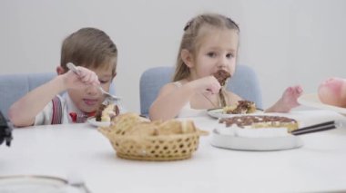 Pasta yiyen beyaz bir çocukla bir kızın yakın çekim yüzleri. Küçük erkek ve kız kardeş doğum günü partisinde tatlı yemeğin tadını çıkarıyorlar. Mutluluk, rahatlık..