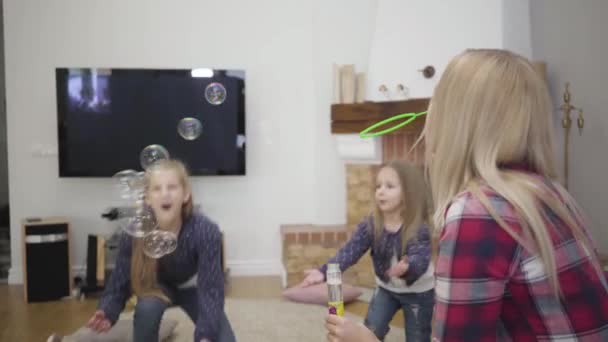 Back view of blond Caucasian woman blowing soap bubbles for daughters. Cheerful girls laughing and trying to catch bubbles. Happy family resting at home. Happiness, leisure, unity. — Stock Video
