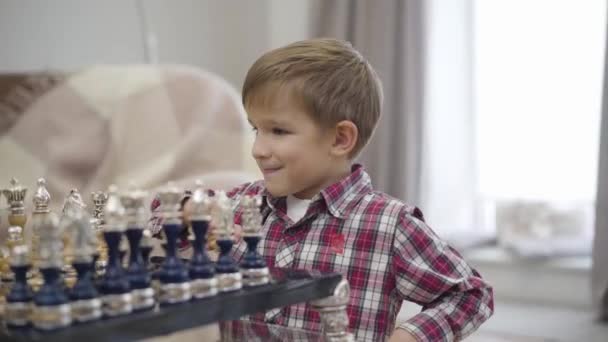Közelkép egy cuki fehér fiúról, aki a sakktábla előtt ül és mosolyog. Okos gyerek, élvezi a hobbiját idebent. Intelligencia, intellektus, gyermekkor.
