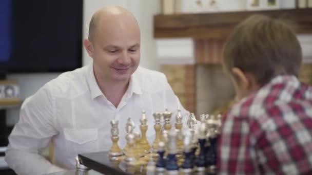 Portrait d'un homme caucasien heureux jouant aux échecs avec son fils. Un père intelligent souriant éduquant son enfant à la maison. Famille, intellect, unité . — Video