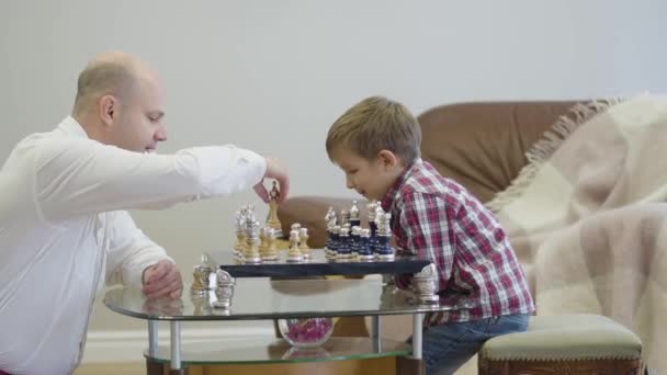 チェス盤の前に座って賢い息子と遊ぶ白いシャツの白人男性のサイドビュー。かわいい賢い少年は家で父と無料の食事を過ごす。団結、知性、教育. — ストック動画