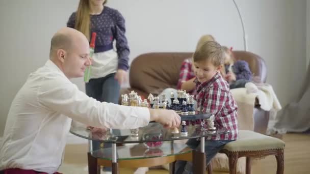 Ung kaukasisk far spelar schack med sonen som äldre dotter på gång. Mor och yngre flicka sitter på soffan i bakgrunden och läser. Positiva familjespenderande helger hemma. — Stockvideo
