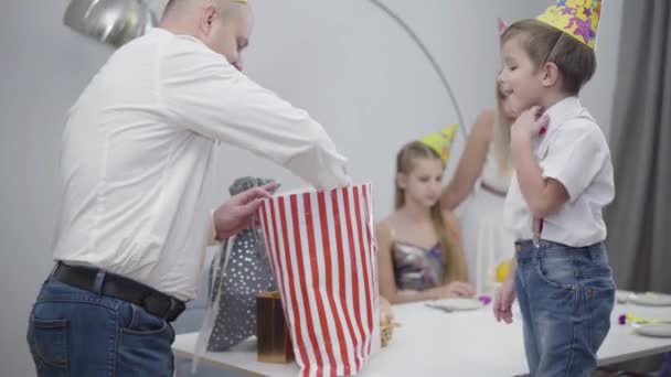 Portret van een volwassen blanke man met feestmuts die zijn zoon een speelgoedauto geeft als verjaardagscadeau. Gelukkig vader neemt speelgoed mee en geeft het aan een gelukkig jongetje. Feesten, feesten, eenheid. — Stockvideo