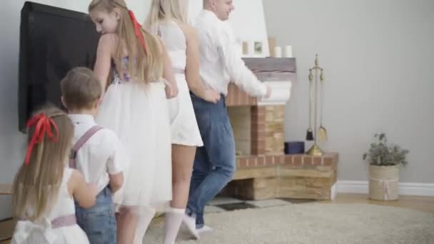 Wesoła biała rodzina tańcząca wokół stołu w domu. Uśmiechnięty ojciec, matka i trójka dzieci trzymających się i poruszających. Wypoczynek, imprezowanie, zabawa. — Wideo stockowe