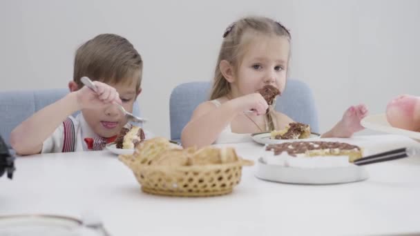 Arcok cuki fehér fiúkról és lányokról, akik tortát esznek. A kistestvérek édességet esznek a szülinapi bulin. Boldogság, szabadidő.. — Stock videók