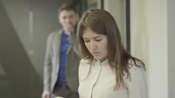 Nahaufnahme einer schönen jungen kaukasischen Frau, die Dokumente durchsieht, während ein gutaussehender Mann im Hintergrund seinen Anzug überprüft und auftaucht. Büro-Romantik, Flirt, Flirt. — Stockvideo