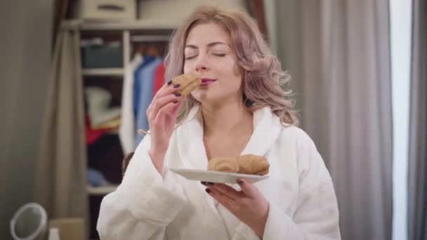 Retrato de una hermosa mujer caucásica en albornoz blanco oliendo sabroso croissant y mordiéndolo. Encantadora dama disfrutando de la comida dulce en casa por la mañana. Dieta, belleza, estilo de vida . — Vídeo de stock