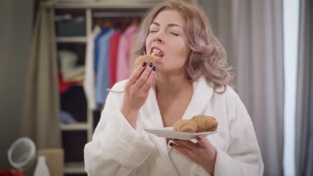 Retrato de hambrienta hermosa chica caucásica disfrutando de comer croissant. Mujer joven masticando sabroso bollo en casa. Dieta, comida, nutrición . — Vídeos de Stock