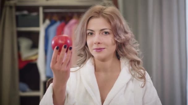 Портрет чарівної кавказької дівчини, що кусає червоне яблуко і показує великий палець вгору. Прекрасна молода жінка їсть здорову їжу вдома. Їжа, харчування, спосіб життя. — стокове відео