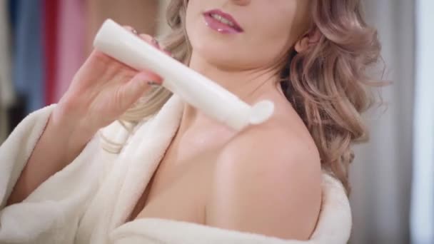 Close-up van charmante blanke vrouw die crème op de schouders aanbrengt. Mooie dame in witte badjas zorgt thuis voor haar huid. Wellness, schoonheid, levensstijl. — Stockvideo