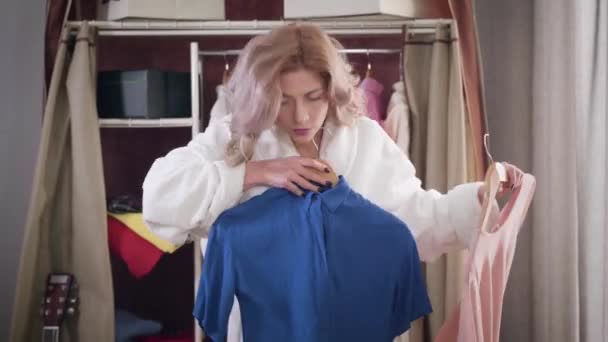 Portrait de jeune fille confiante essayant chemise bleue et chemisier rose. Belle femme en peignoir blanc choisissant une tenue élégante le matin. Style, mode . — Video