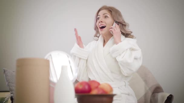 Retrato de la alegre chica caucásica en albornoz blanco sentado en el sofá y hablando por teléfono. Hermosa mujer haciendo gestos emocionales. Felicidad, estilo de vida, ocio . — Vídeos de Stock