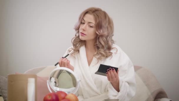Porträtt av ung vit kvinna som tittar på spegeln och applicerar ögonskuggor. Charmig tjej i vit badrock gör sig redo på morgonen. Kosmetika, make-up, skönhet. — Stockvideo