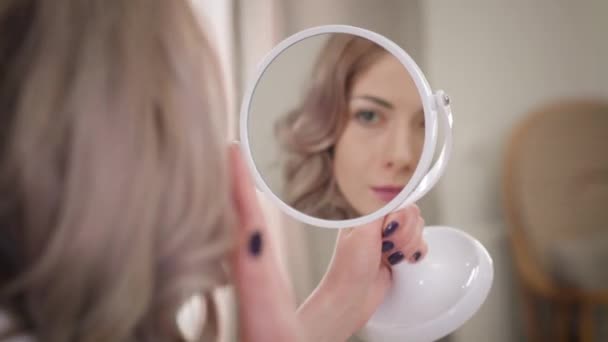 Розмите відображення посміхається кавказькій дівчині в маленькому круглому дзеркалі. Мила жінка милується собою і чіпляється за волосся. Краса, спосіб життя. — стокове відео