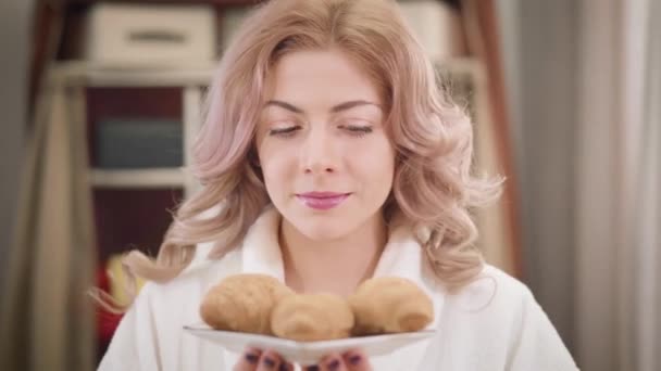 Retrato de una hermosa chica caucásica oliendo sabrosos cruasanes y mostrando el plato a la cámara. Mujer joven feliz con ojos de avellana y pelo rizado proponiendo dulces. Dieta saludable, estilo de vida . — Vídeos de Stock