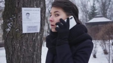 Heyecanlı beyaz kadın portresi telefonla arıyor ve ağaçta asılı olan kayıp çocuk ilanına bakıyor. Empatik kız kayıp kişiyi bulmak için insanlara yardım ediyor. Adam kaçırma, kayıp, arama.