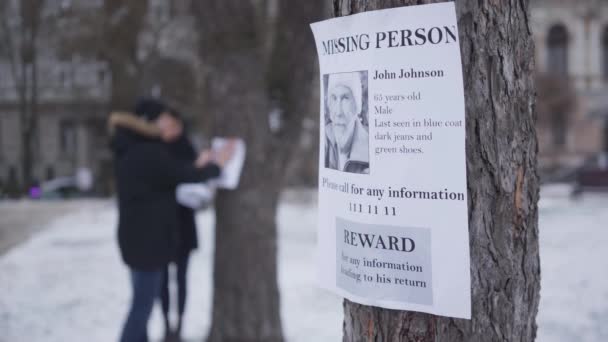 Close-up of missing person ad with information about lost senior Caucasian man. Blurred people hanging ads on trees at the background. Family searching for their grandfather. Unity, loss, kidnapping. — 비디오