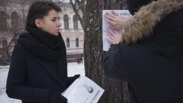 Běloch věší na strom reklamu na pohřešovaného syna jako rozrušenou manželku, která stojí s ostatními brožurkami. Lidé odcházejí ze záběru. Únos, ztráta, zoufalství. — Stock video