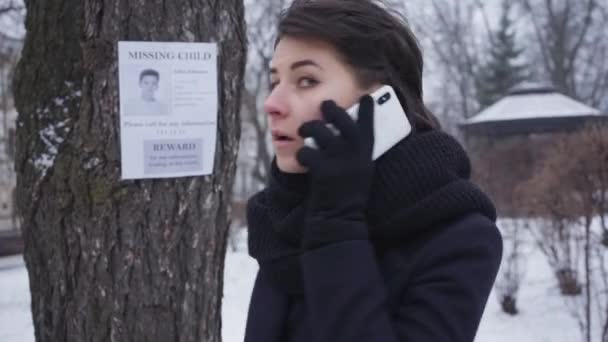 Portret podekscytowanej młodej kobiety dzwoniącej przez telefon i patrzącej wstecz na ogłoszenie o zaginionym dziecku wiszące na drzewie. Empatyczna dziewczyna pomaga ludziom odnaleźć zagubioną osobę. Porwanie, utrata, przeszukanie. — Wideo stockowe
