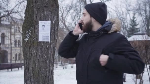 Egy szakállas, fiatal, fehér férfi portréja, aki eltűnt gyerekreklám mellett áll és telefonál. Jóképű fickó, aki információt szolgáltat az eltűnt személyről a rokonainak. Emberrablás, házkutatás.. — Stock videók