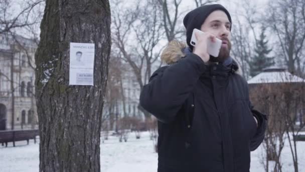 Homem caucasiano barbudo jovem falando ao telefone e olhando para a pessoa desaparecida anúncio pendurado na árvore. Cara empática em roupas de inverno ajudando as pessoas a encontrar pessoa perdida. Rapto, perda, busca . — Vídeo de Stock