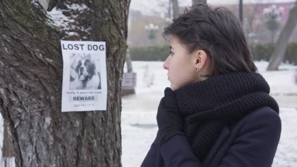 Retrato de cerca de la triste chica caucásica parada en la calle de invierno y mirando a su alrededor. Anuncio sobre el perro perdido colgado en el árbol al fondo. Pérdida, problema, desesperación . — Vídeos de Stock