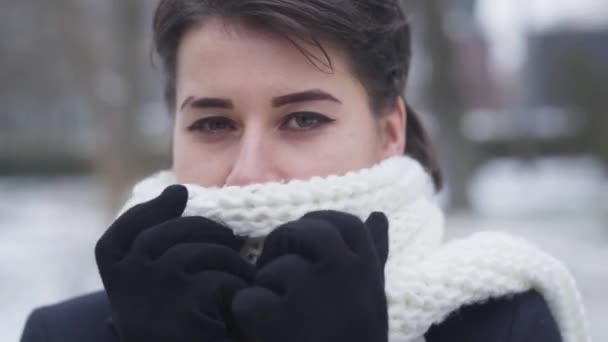 Gros plan de la jeune fille brune caucasienne avec les yeux bruns cachant visage en écharpe chaude blanche. Portrait de femme mignonne regardant la caméra comme debout sur la rue d'hiver. Loisirs, beauté . — Video