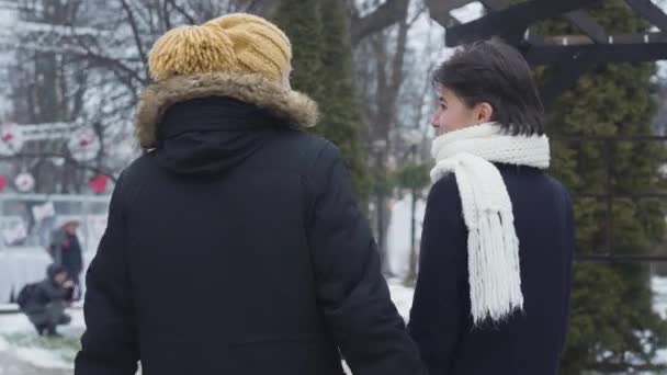 Vista posteriore di felice giovane coppia caucasica passeggiando lungo la strada invernale e parlando. ragazzo barbuto in cappello giallo e affascinante ragazza in sciarpa bianca incontri all'aperto. Amore, svago, felicità . — Video Stock