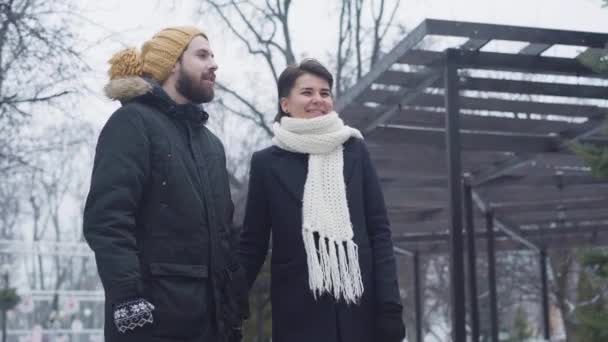 Střední záběr usmívajícího se bělošského páru procházejícího se venku. Muž v legračním žlutém klobouku a okouzlující žena v bílém šátku datování v zimě. Láska, vztah, sblížení. — Stock video